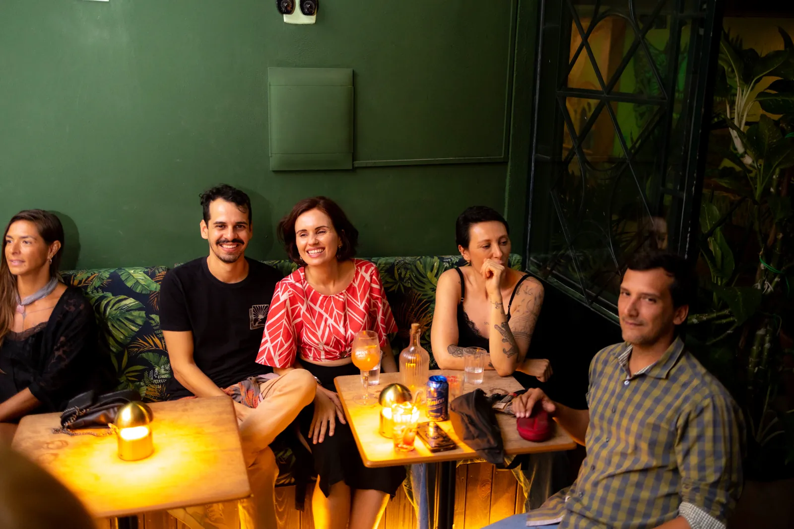 Amigos sorrindo no sofá do Na Moita Bar, com drinks na mesa e a parede verde ao fundo