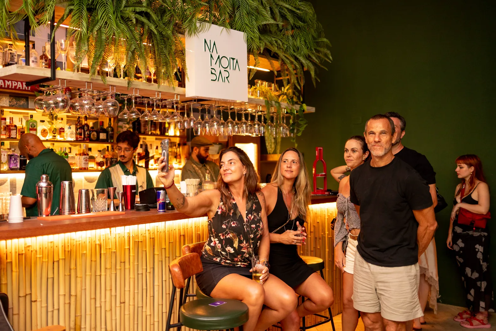 Público no balcão de bambu do Na Moita Bar, sob o letreiro e a cristaleira iluminada