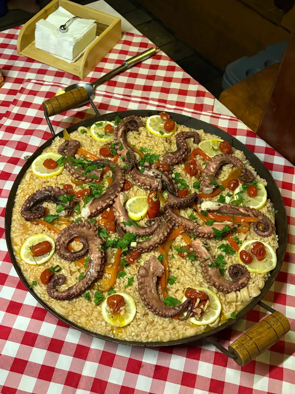 Travessa grande de arroz de polvo com tentáculos, limão e tomatinhos sobre a mesa de toalha xadrez do barco