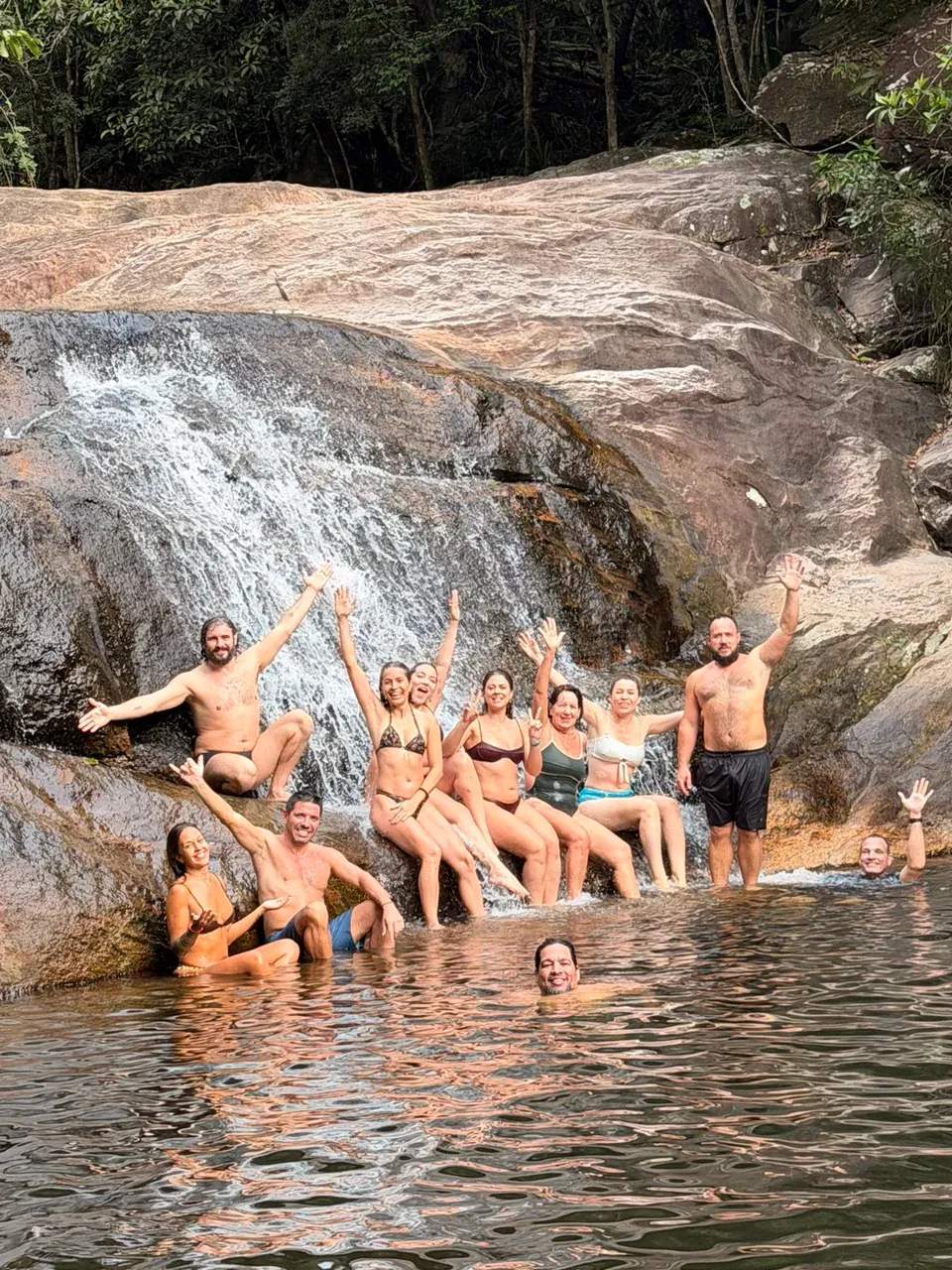 Grupo de hóspedes dentro do poço de uma cachoeira na mata, sentados nas pedras da queda dágua