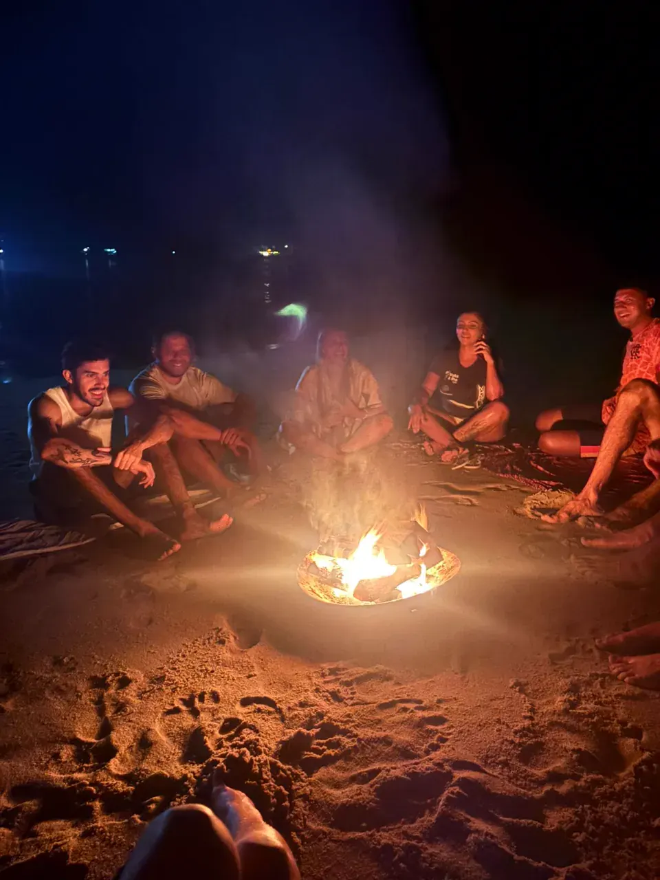 Grupo sentado na areia em volta de uma fogueira à noite, com o mar escuro ao fundo