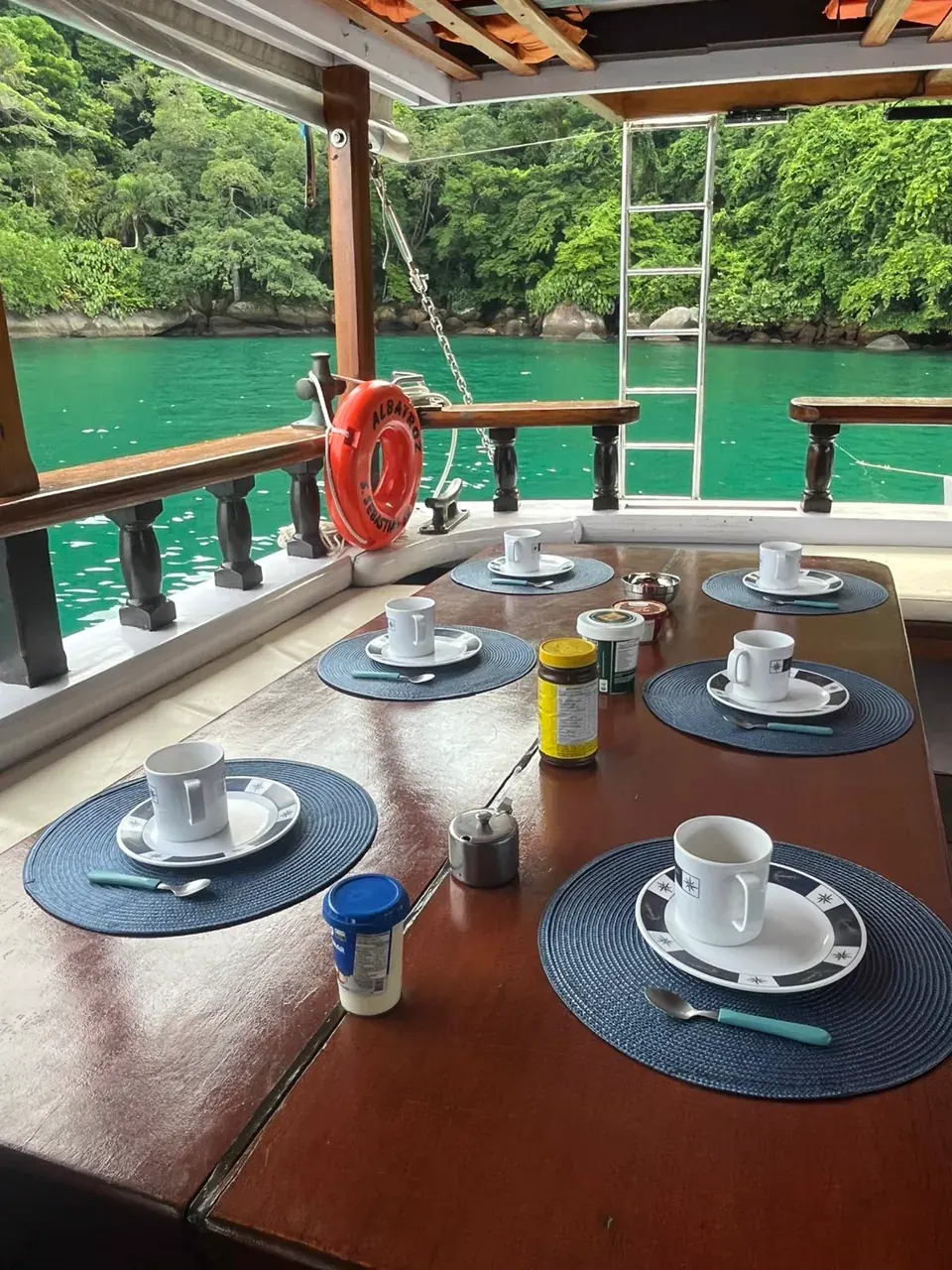 Mesa de café da manhã posta no convés coberto da escuna, com xícaras sobre jogos americanos azuis, boia salva-vidas laranja e a água verde de uma enseada ao fundo