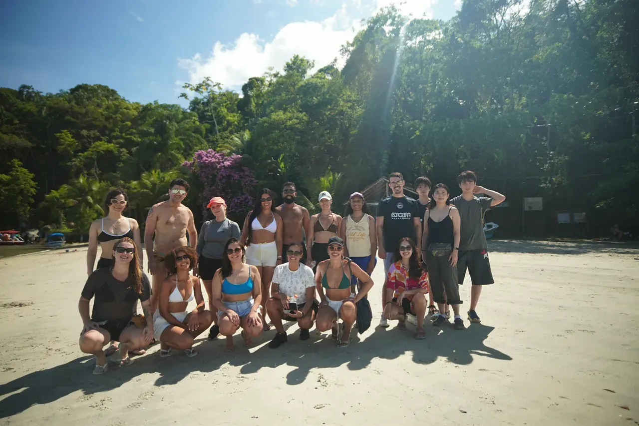 Grupo de hóspedes reunido na areia de uma praia deserta da Cajaíba, com a mata fechada e uma primavera florida ao fundo