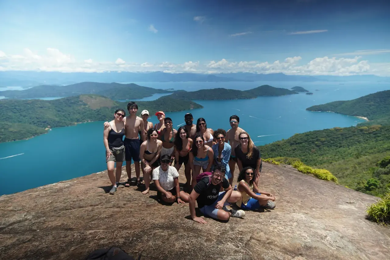 Grupo de hóspedes posando sobre a laje de pedra no alto do Pão de Açúcar do Mamanguá, com o fiorde e as ilhas da baía de Paraty ao fundo