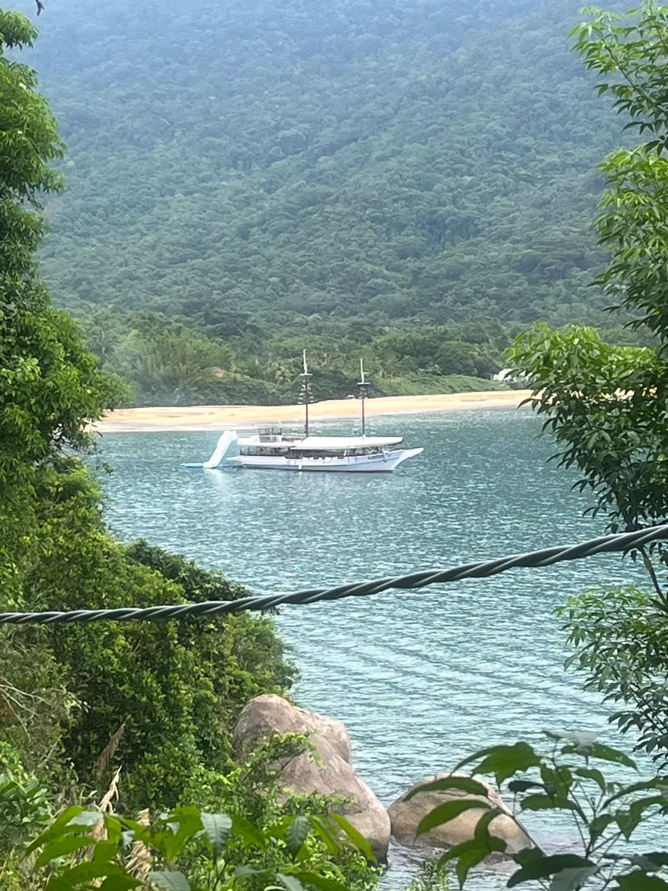 A escuna Albatroz fundeada em uma enseada de água verde cercada de mata, vista de cima da trilha na costeira