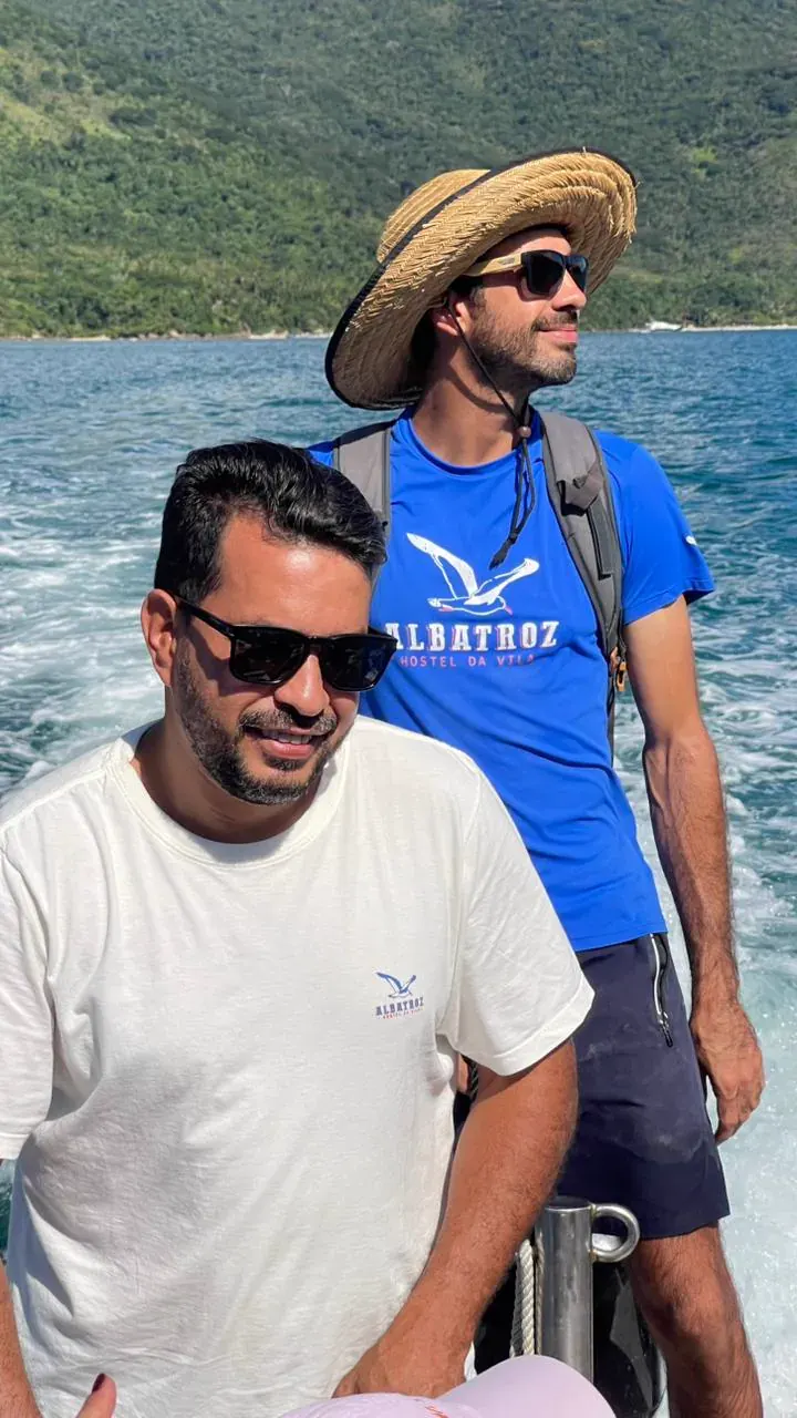 Dois tripulantes de camiseta do Albatroz no bote de apoio, um deles de chapéu de palha, com a costeira de mata ao fundo