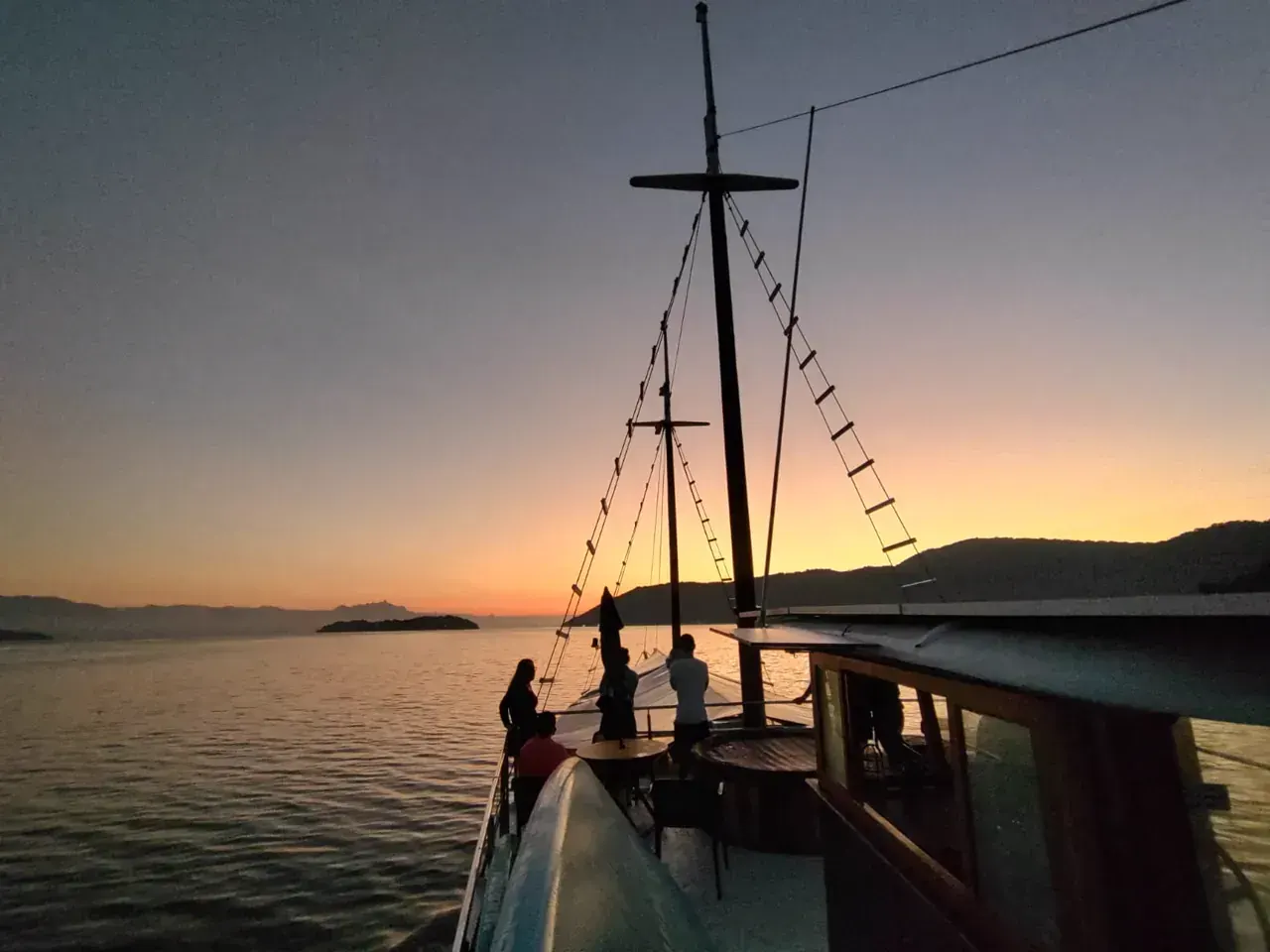 Silhuetas de viajantes no convés do Albatroz ao entardecer, com os mastros contra o céu alaranjado