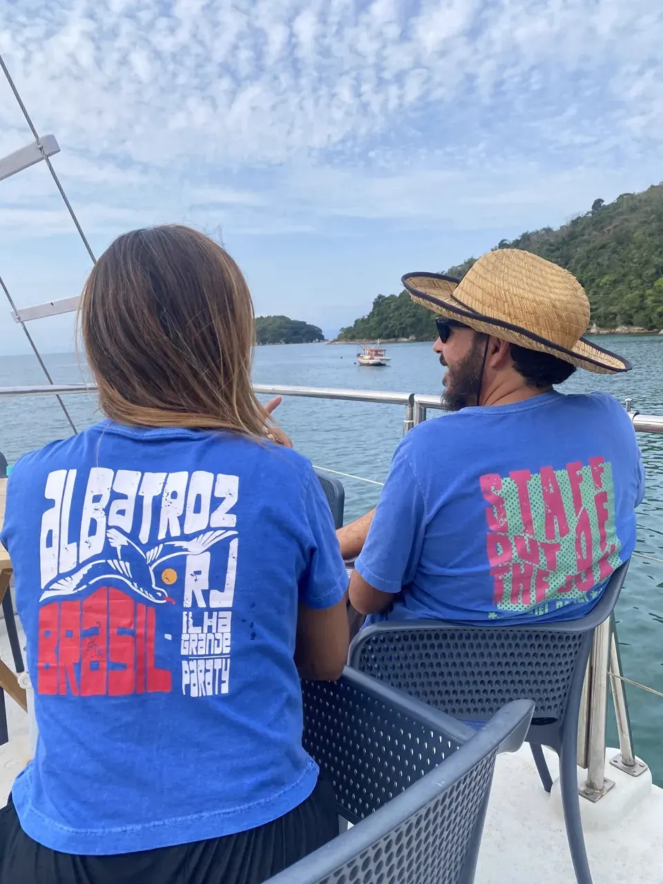 Dois hóspedes de costas no convés, com camisetas azuis do Albatroz, olhando a enseada verde e um barco de pesca fundeado ao longe