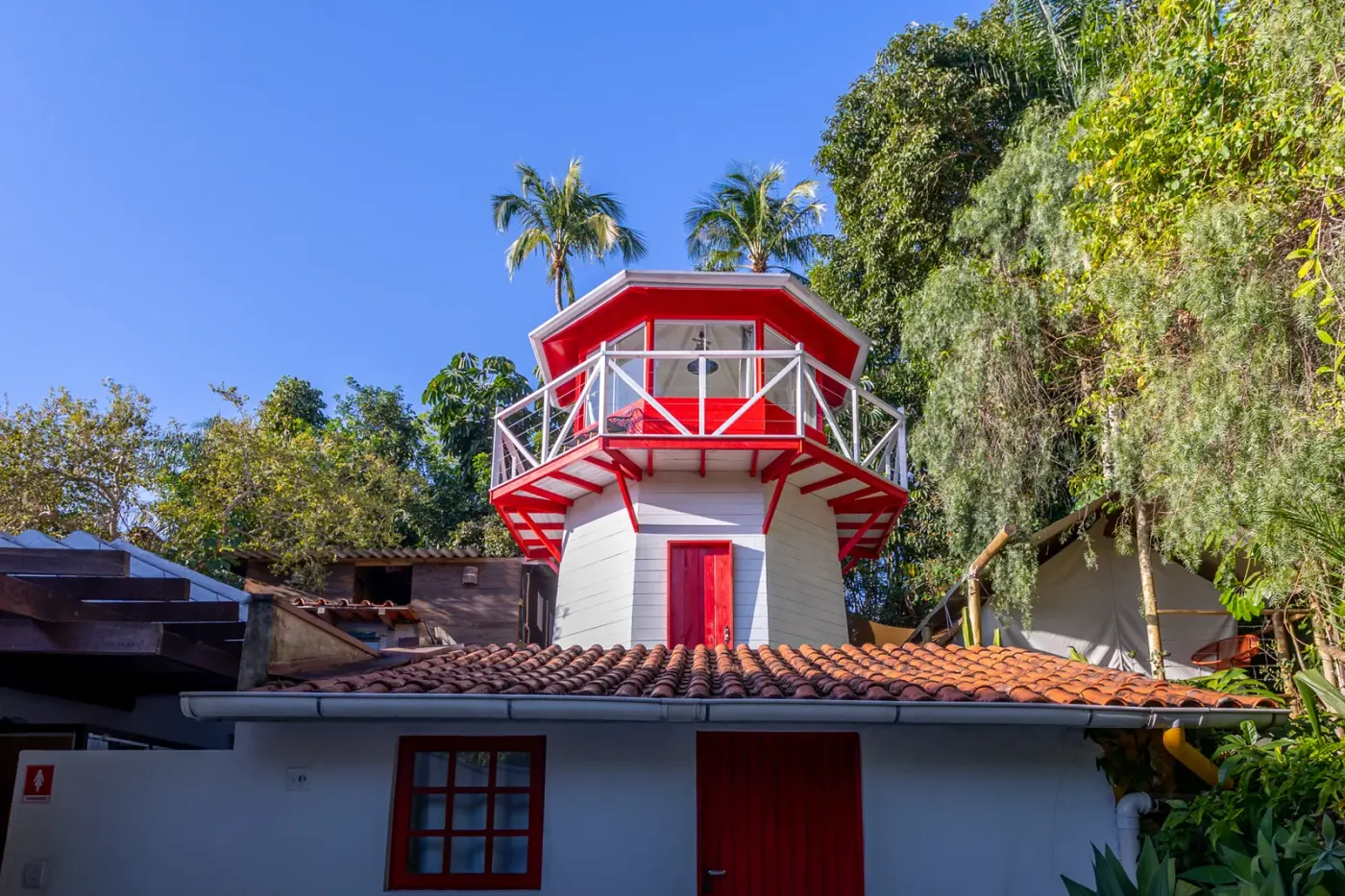 Farol branco e vermelho com mirante no topo, entre palmeiras e telhados de telha