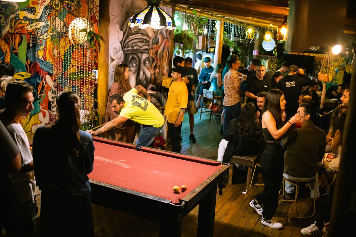Salão de bar com mesa de sinuca vermelha, murais coloridos nas paredes e grupos de pessoas conversando em volta