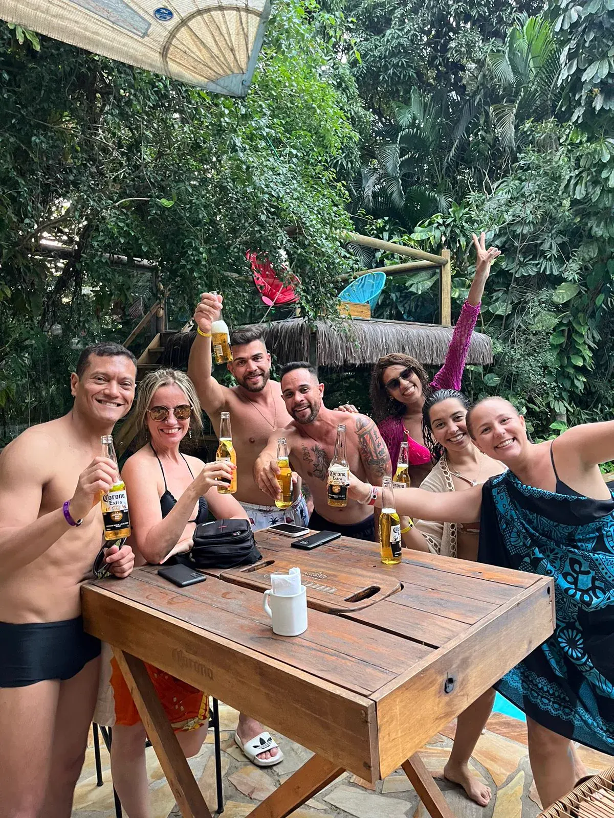 Grupo de hóspedes brindando com garrafas em volta de uma mesa alta de madeira ao lado da piscina, com a mata fechada ao fundo