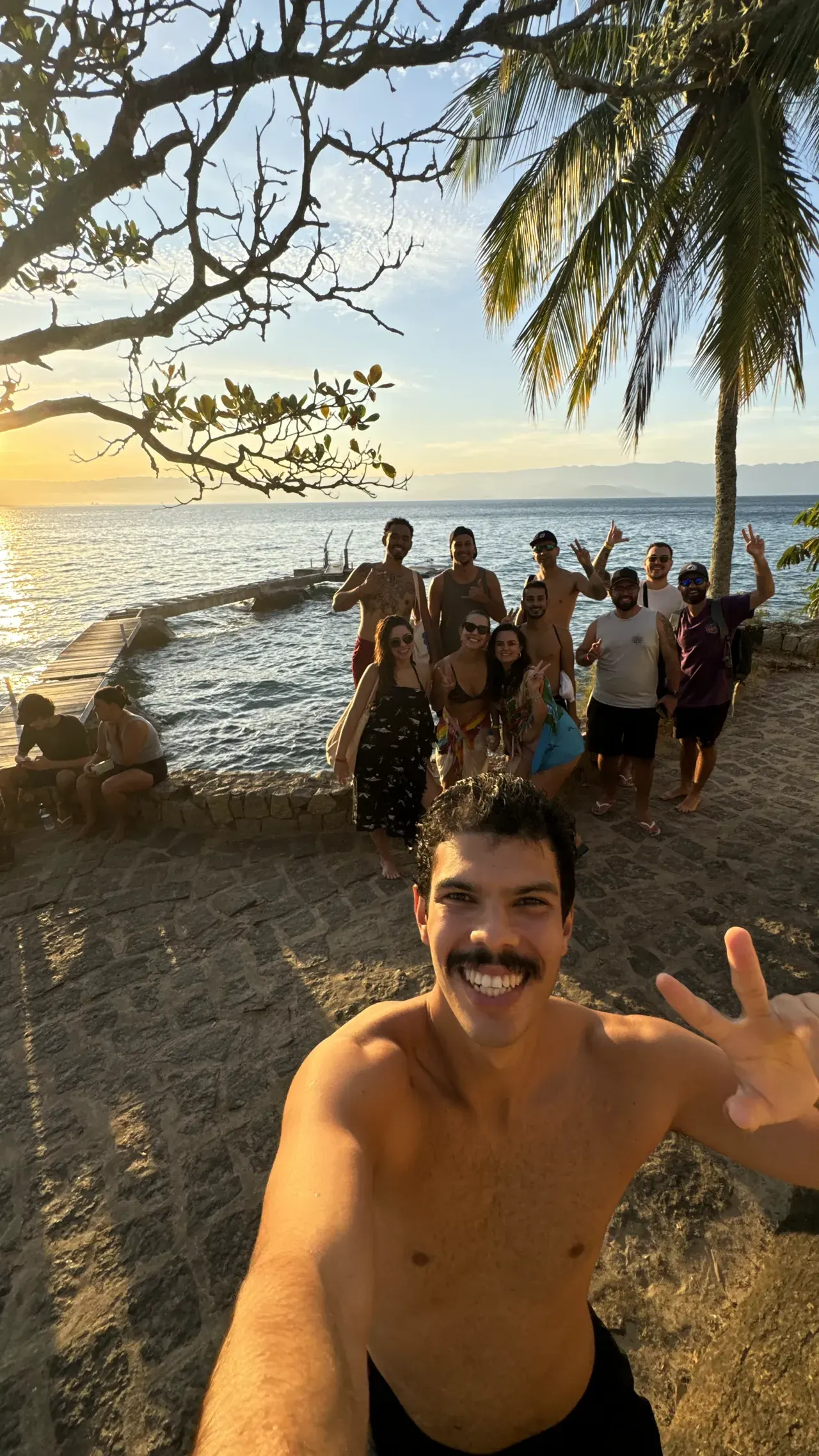 Grupo de viajantes reunido à beira do canal de São Sebastião no pôr do sol, com pier de pedra ao fundo