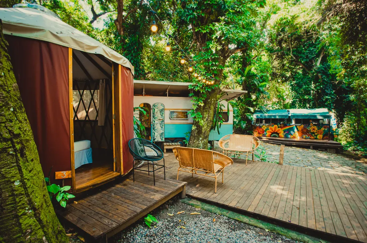 Yurt, trailer e ônibus coloridos entre as árvores, ligados por deck de madeira com varal de luzes