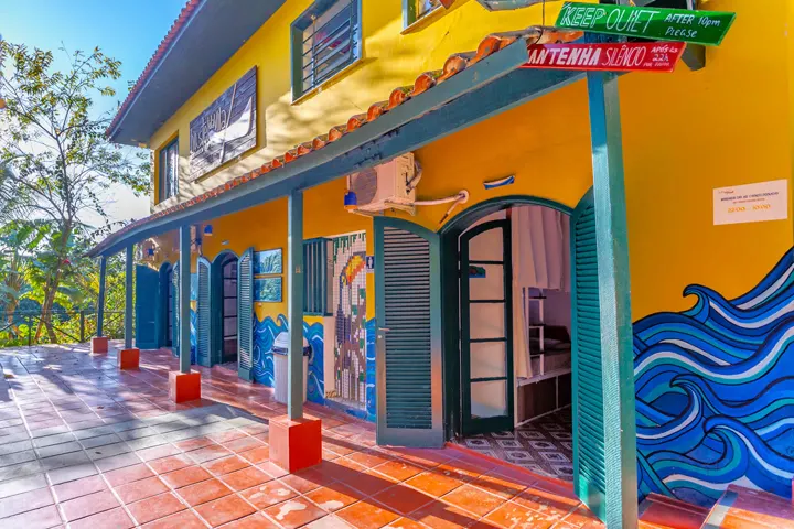 Fachada amarela de hostel com portas verdes, murais de ondas e placa pedindo silêncio depois das 22h