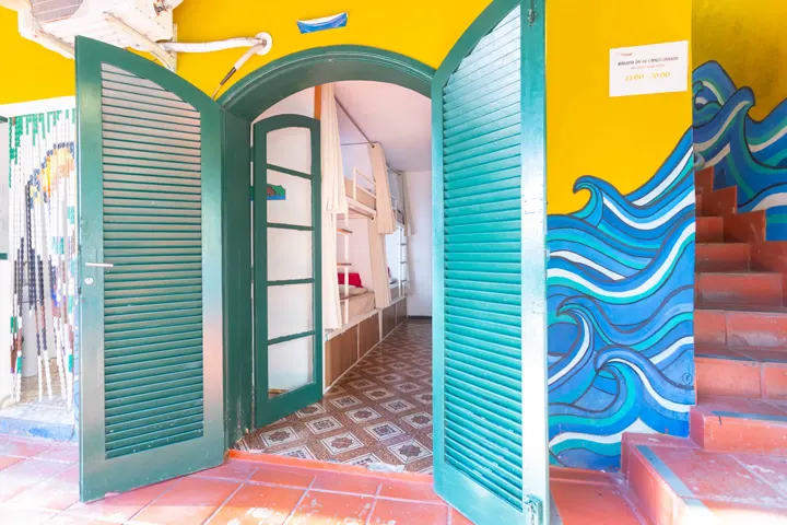 Portas verdes abertas revelando um dormitório com beliches de cortinas brancas e piso de ladrilho antigo