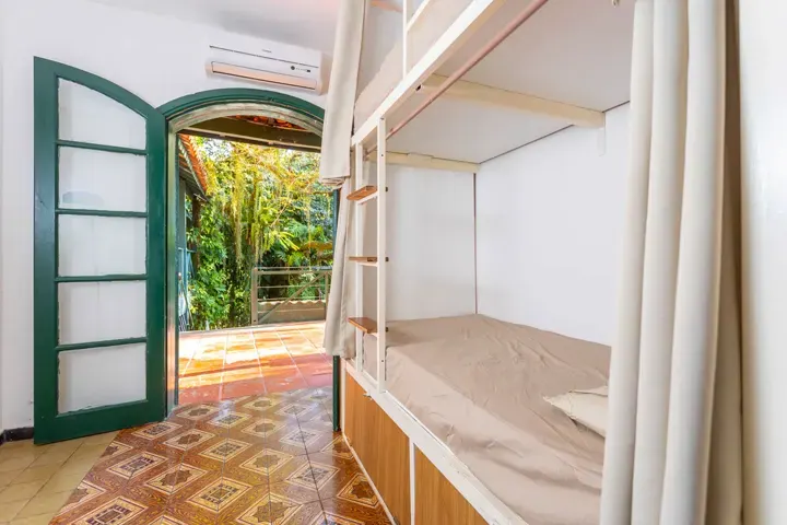 Dormitório claro de hostel com beliche de cortina individual e porta verde aberta pra um terraço com vegetação