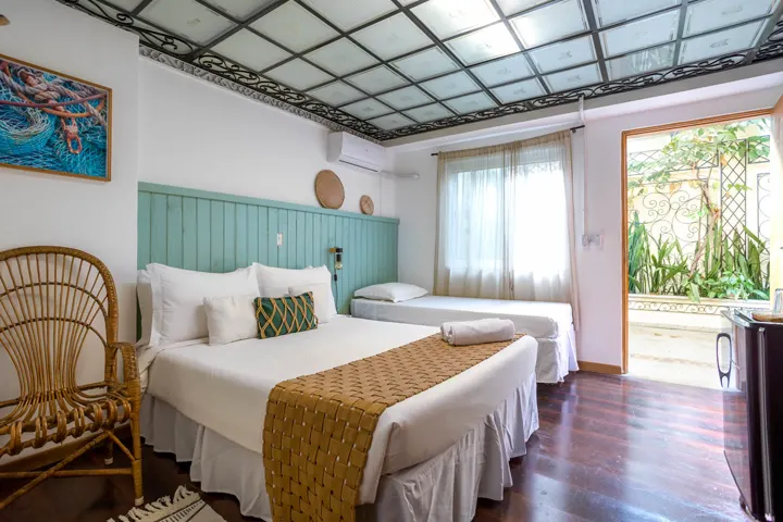 Suíte família do Vilarejo com cama de casal, cama de solteiro ao lado, cabeceira de madeira verde-água e porta abrindo pra um pátio com plantas