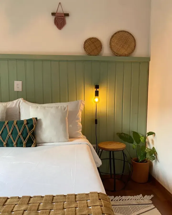 Quarto privativo com cama de casal, lambri verde, luminária acesa e banquinho de madeira