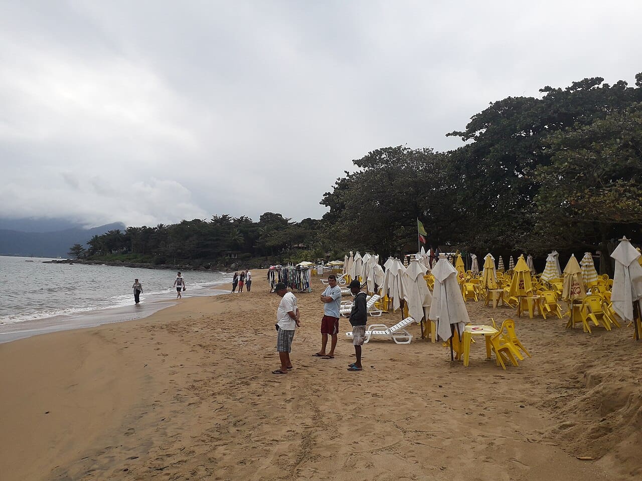 Faixa de areia clara com mar calmo e árvores na Praia do Curral, em Ilhabela